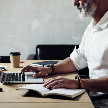 Adult bearded businessman wearing a classic glasses and working at the wood table in modern loft.Stylish middle age man using notebook. Horizontal closeup, blurred background